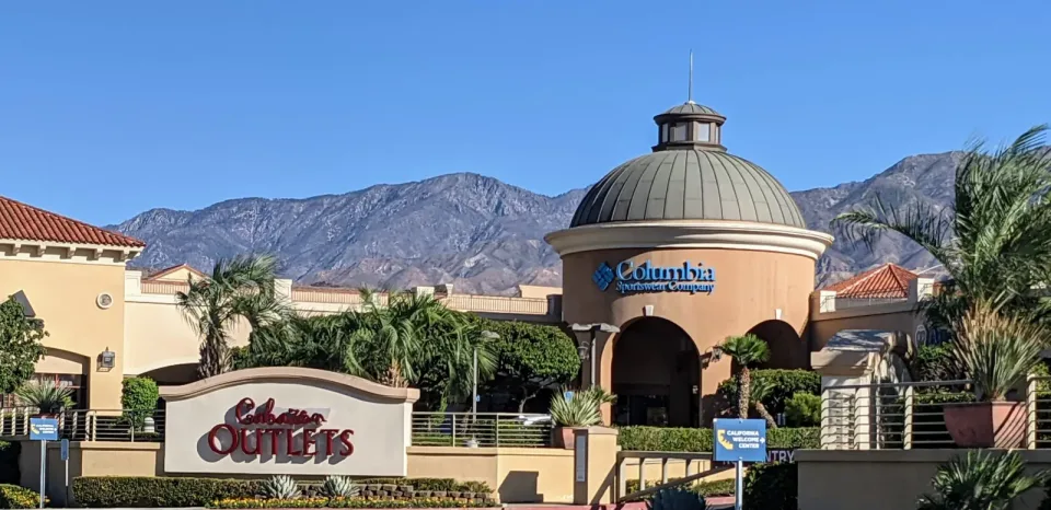 The Cabazon Outlets sign and open-air storefronts sit beneath palms and the San Gorgonio Pass mountains.