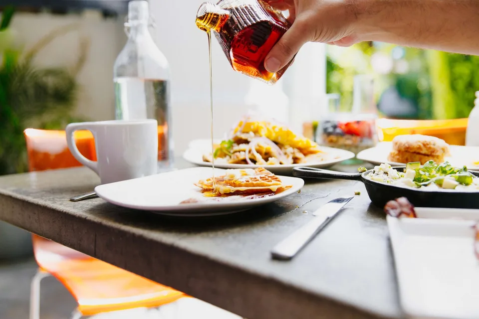 Maple syrup is poured over a waffle at a sunlit breakfast table at Cheeky’s.