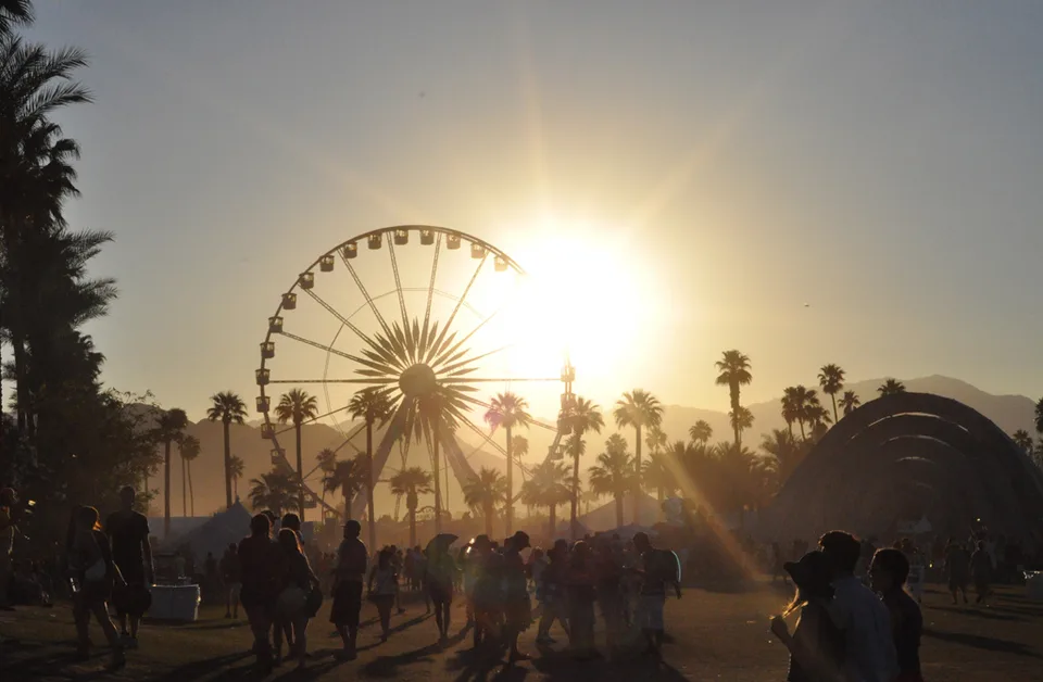 Festivalgoers walk beneath the Coachella Ferris wheel as the sun sets behind palms and mountains.