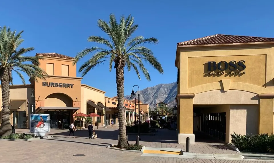 Palm-lined storefronts at Desert Hills Premium Outlets frame the surrounding mountains beneath a clear blue sky.