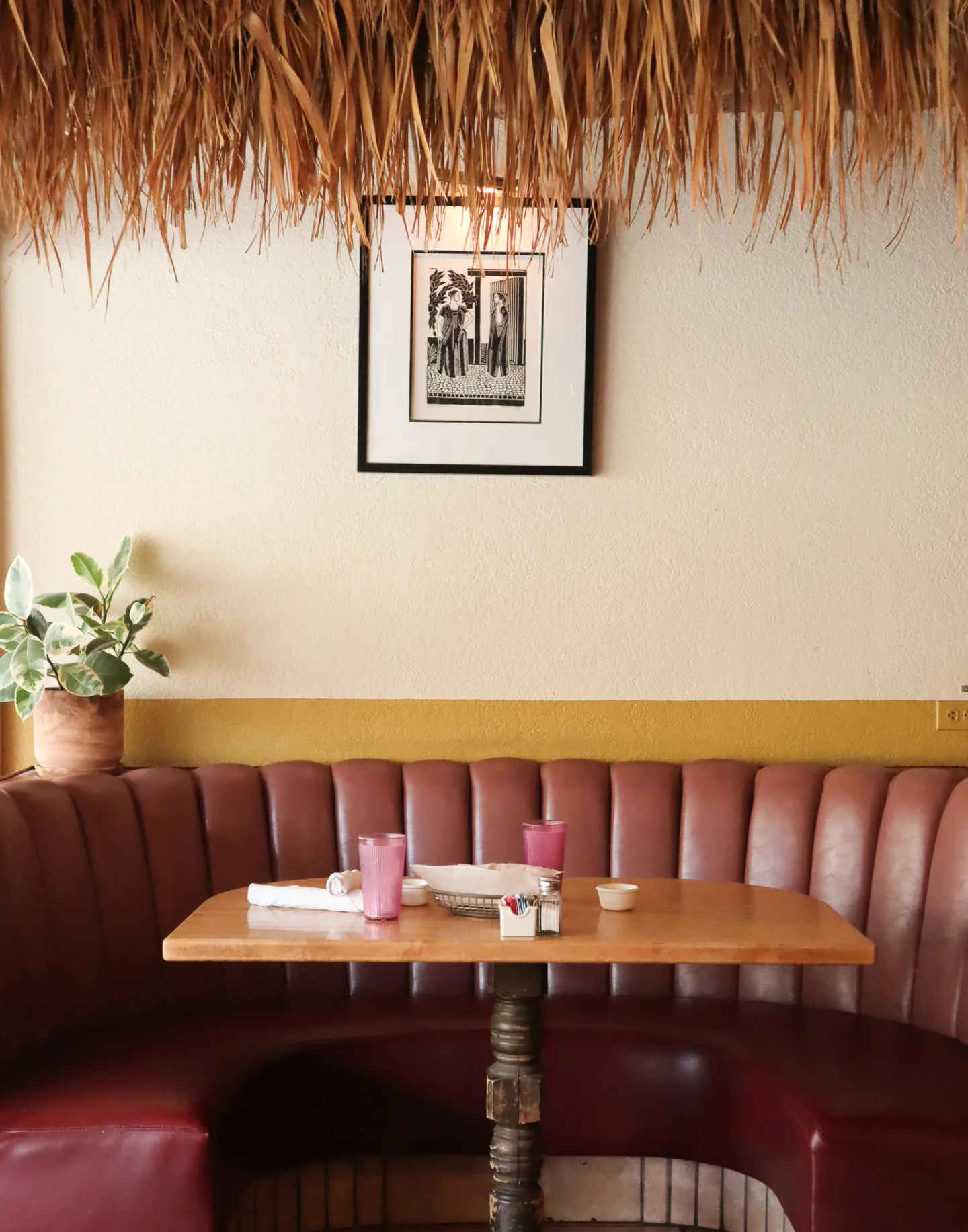 A red leather banquette and wooden table sit beneath framed artwork and a thatched canopy at El Mirasol.