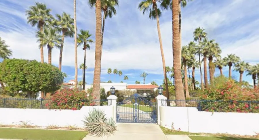 Front gate of the Gene Autry Residence framed by palms, flowering hedges, white walls, and mountains.