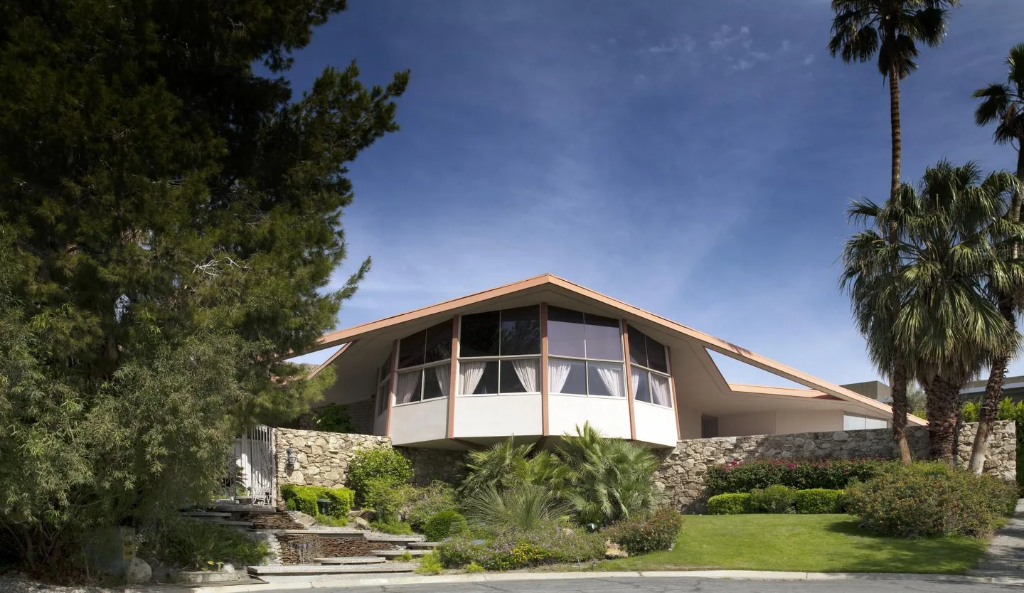 Street-facing view of the House of Tomorrow with its angular roof, glass walls, stonework, palms, and garden.