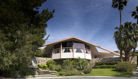 Street-facing view of the House of Tomorrow with its angular roof, glass walls, stonework, palms, and garden.