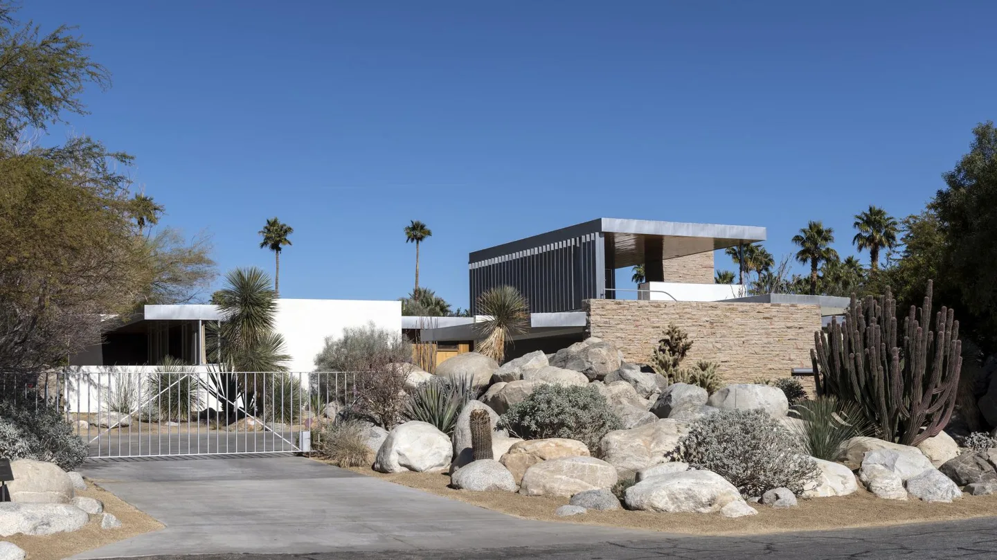 Street-facing view of the Kaufmann Desert House with pale stone, metal shade fins, boulders, cacti, and palms.