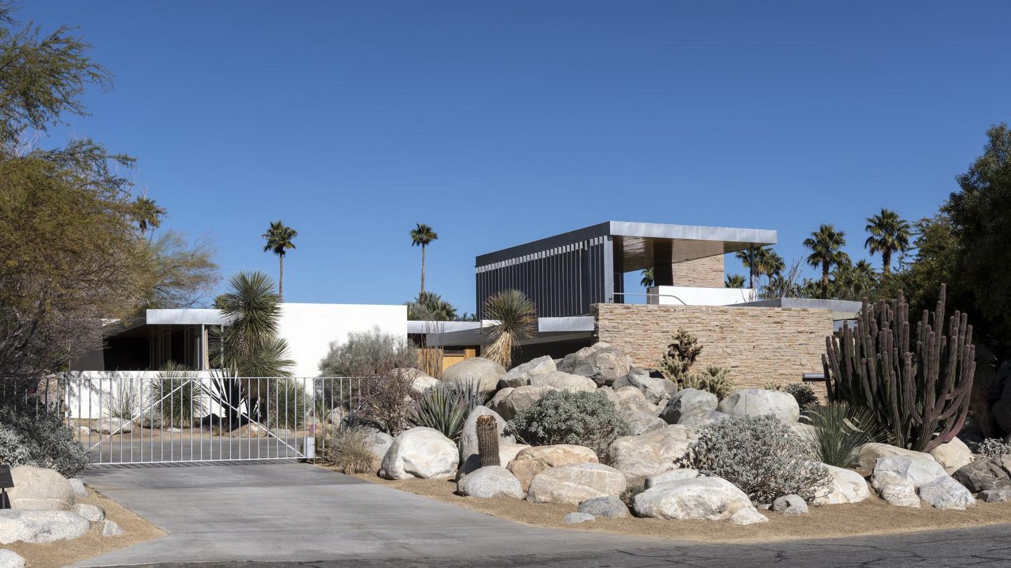 Street-facing view of the Kaufmann Desert House with pale stone, metal shade fins, boulders, cacti, and palms.
