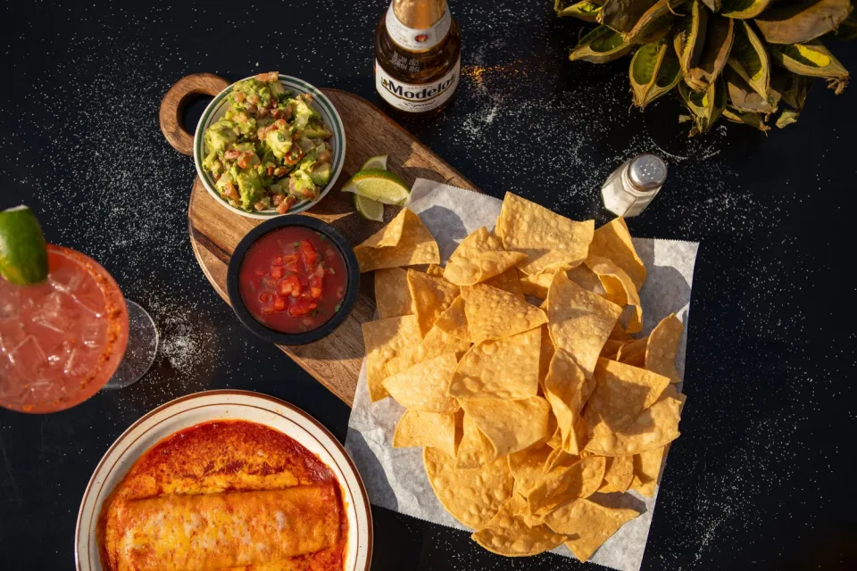 Chips, guacamole, salsa, enchiladas, and drinks are arranged across a dark table at Las Casuelas Original.