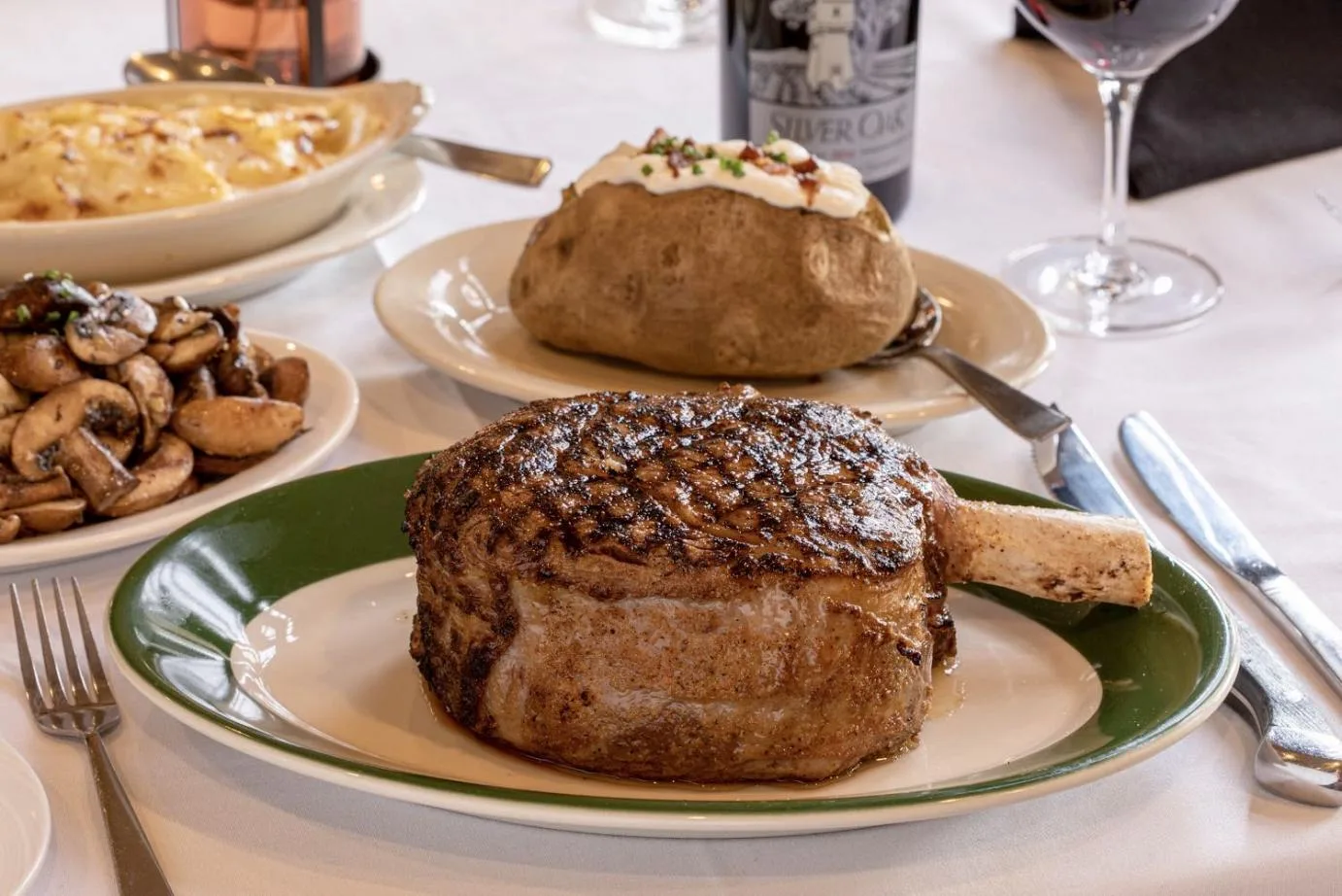 A bone-in steak sits on a green-rimmed plate with a baked potato and sautéed mushrooms.