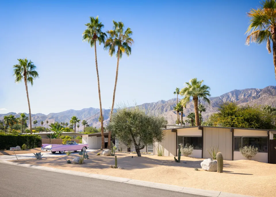 A pink vintage convertible parked outside a low-slung midcentury Palm Springs house, with tall palms and the San Jacinto Mountains beyond.