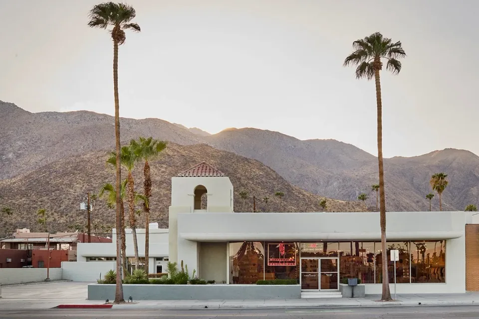 Mojave Flea Trading Post's white Palm Springs storefront stands beneath palms with the San Jacinto Mountains behind it.
