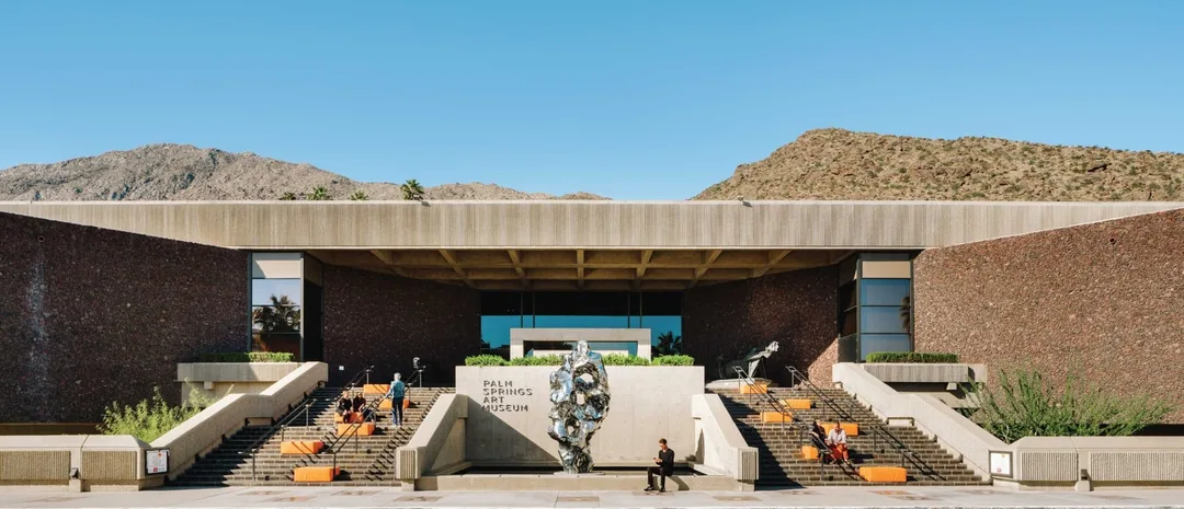 The Palm Springs Art Museum entrance, broad steps, and a reflective metal sculpture sit beneath the San Jacinto Mountains.