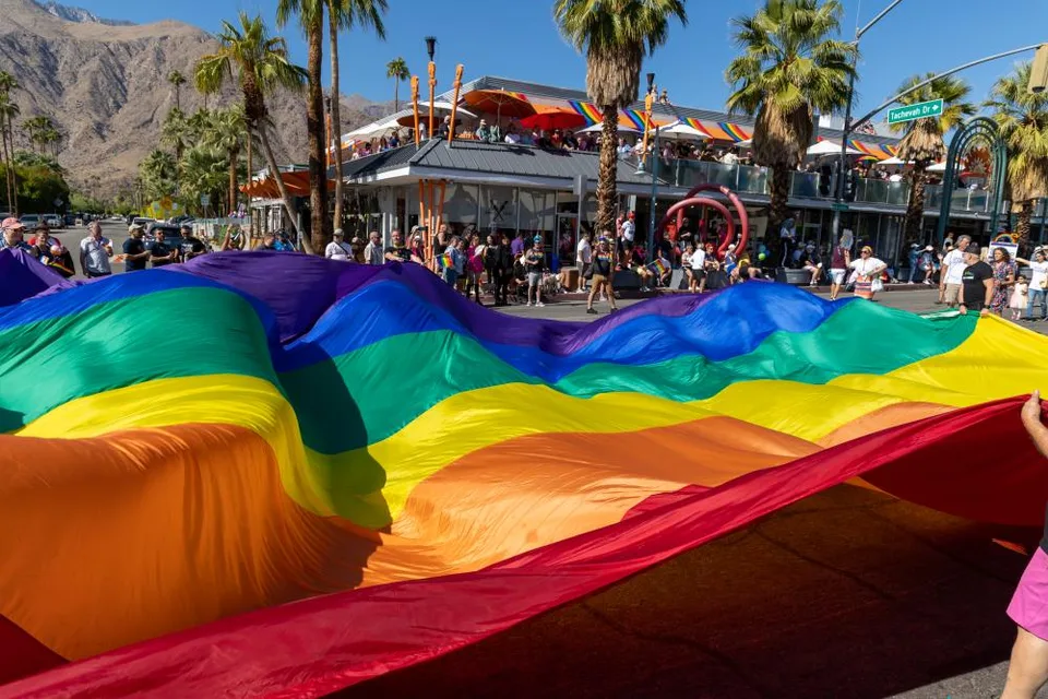 People carry a large rainbow flag along Palm Canyon Drive during Palm Springs Pride, with spectators, palm trees, and the San Jacinto Mountains behind them.