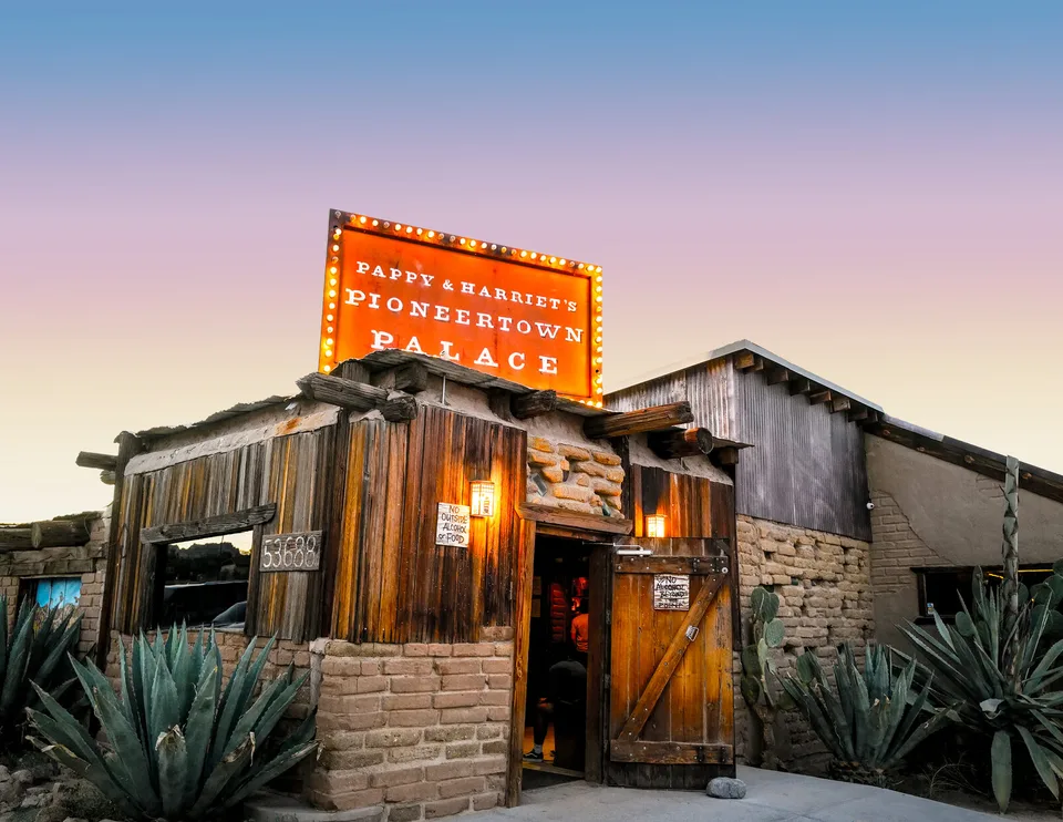 The illuminated Pappy & Harriet’s Pioneertown Palace sign rises above the rustic adobe-and-wood entrance at dusk.