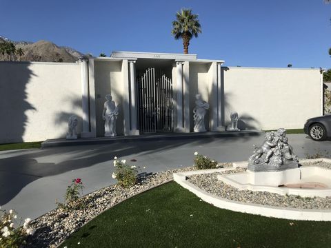White-walled entrance court of Piazza di Liberace with a metal gate, statues, a fountain, trimmed lawn, and the mountain behind.