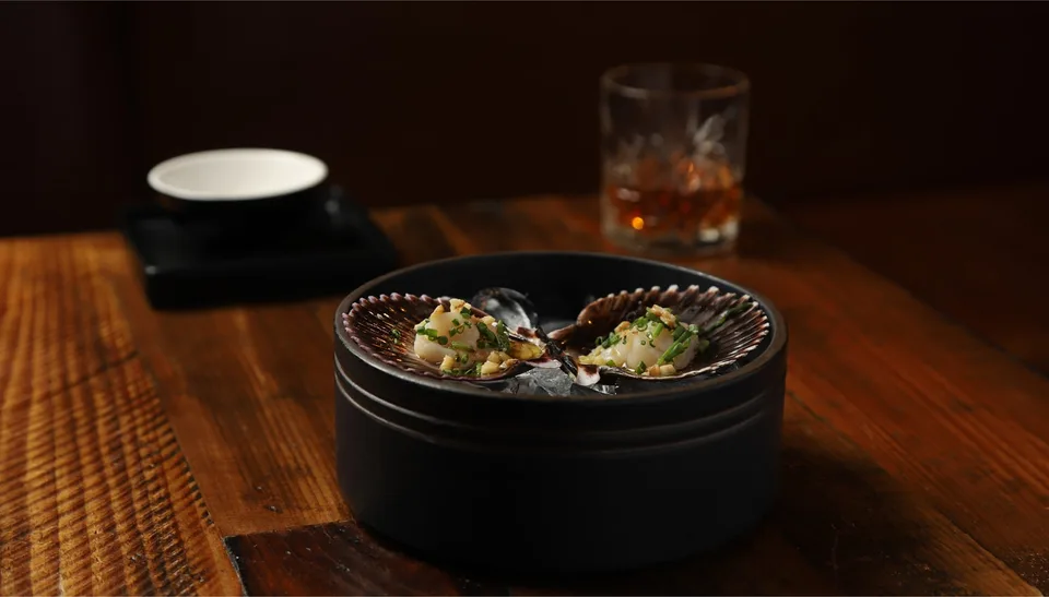 Two scallops in purple shells are served over ice beside an amber drink on a wooden table.