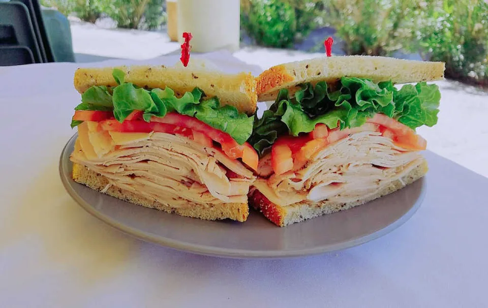 A tall turkey sandwich with cheese, lettuce, and tomato is cut in half on a plate at Sherman’s.