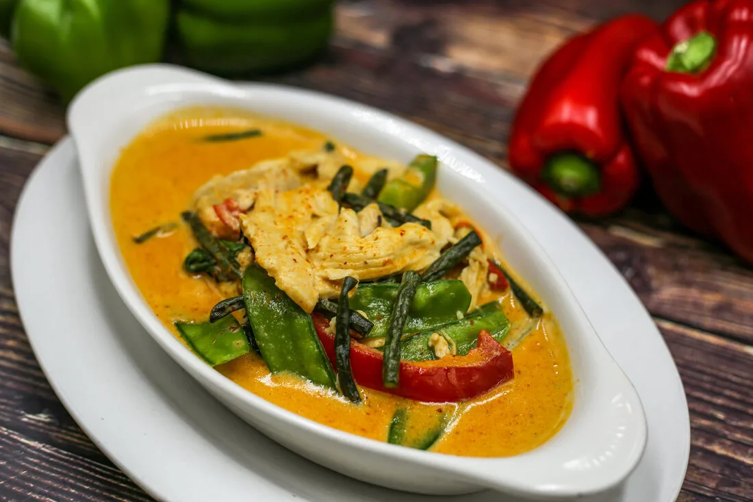Penang curry with sliced chicken, snow peas, green beans, and red bell pepper in a white serving dish.