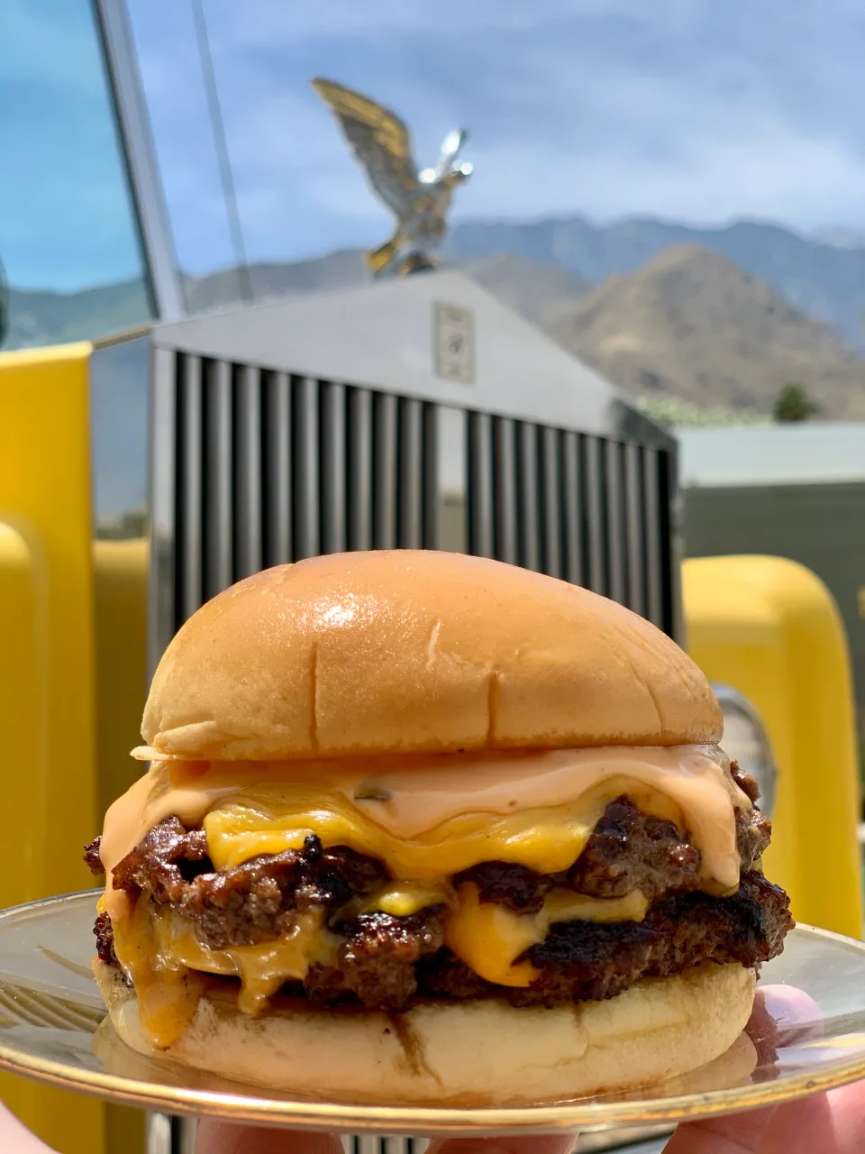 A double smash cheeseburger from The Heyday on a plate, with a vintage car and desert mountains behind it.
