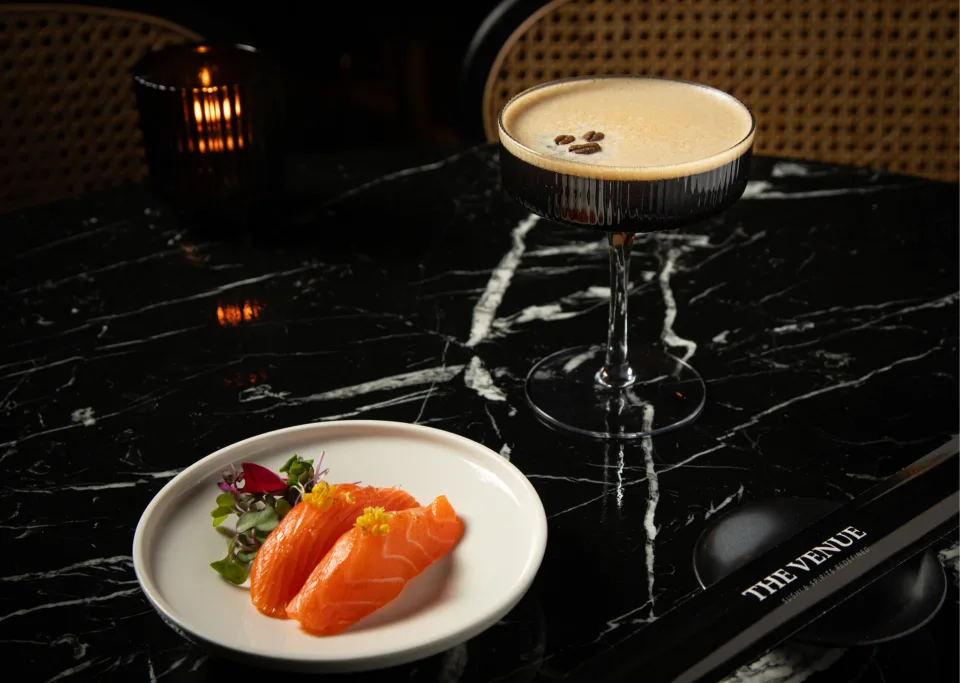 Salmon nigiri and a dark cocktail topped with coffee beans sit on a black marble table beside The Venue-branded chopsticks.