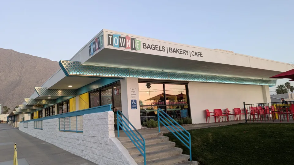 The Townie Bagels storefront has turquoise trim, red patio chairs, and mountains behind it.