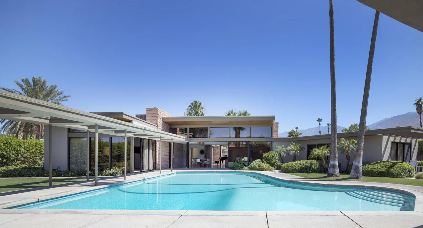 Twin Palms Estate's piano-shaped swimming pool in front of the glass-walled house and two tall palms.