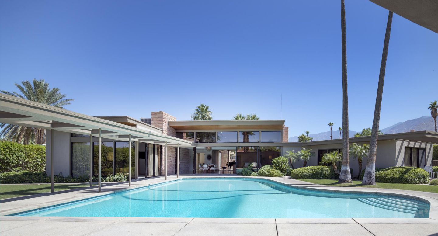 Twin Palms Estate's piano-shaped swimming pool in front of the glass-walled house and two tall palms.