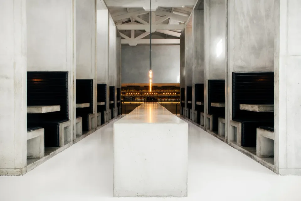 A long white communal table leads between concrete booths toward the illuminated bar at Workshop.